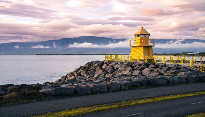 A small, bright yellow lighthouse stands on a rocky pier next to the ocean, with a backdrop of distant mountains under a dramatic, purple-hued cloudy sky.