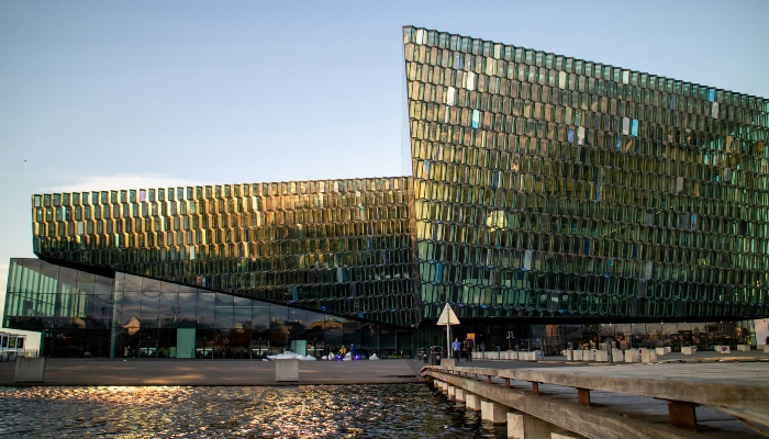 The modern glass facade of the Harpa Concert Hall in Reykjavik, with its geometric panels reflecting the golden light of the sun, viewed from the waterfront.