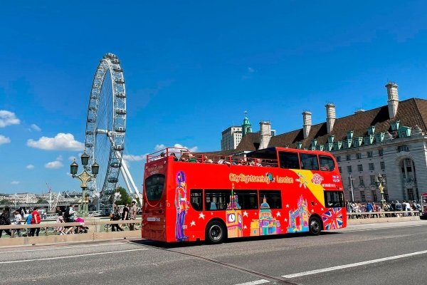 Red City Sightseeing London bus passing the London Eye on a sunny day, part of the city’s most booked sightseeing bus experiences in London.