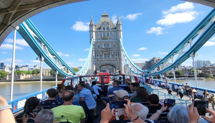 Open-top sightseeing bus crossing Tower Bridge with passengers photographing the view, one of the most booked sightseeing bus experiences in London.