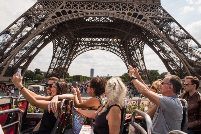 Sightseers on the upper deck of a hop-on hop-off bus taking photos beneath the Eiffel Tower during a Tootbus Paris tour.
