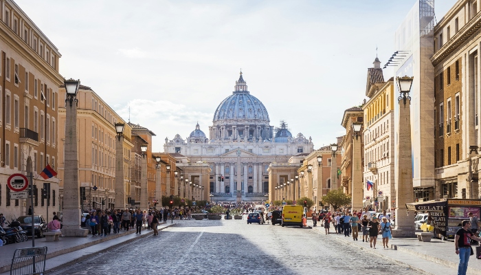 View of St. Peter’s Basilica from Via della Conciliazione with crowds walking toward Vatican City on a bright day.