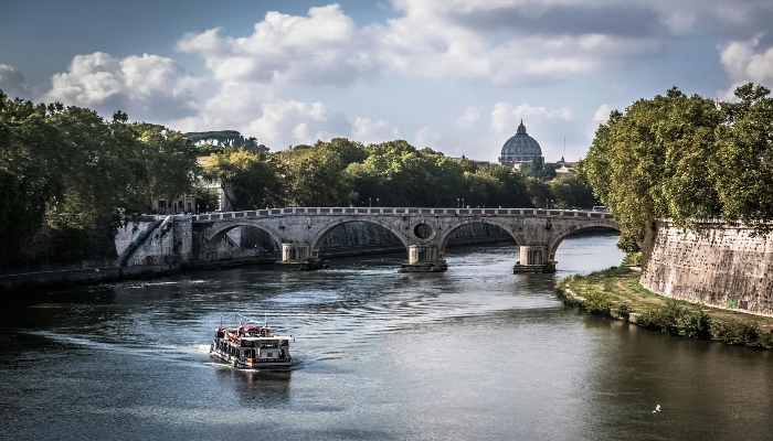 Boat cruising along the Tiber River with a historic Roman bridge and St. Peter’s Basilica in the background during daylight.