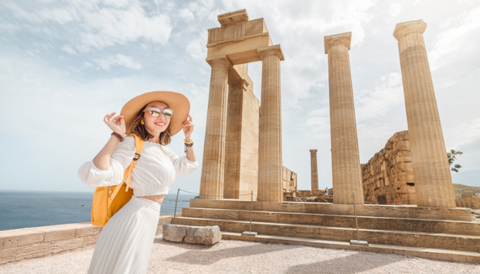 Female traveler at ancient Greek temple ruins in Athens with sea view, sightseeing historic landmarks in summer.