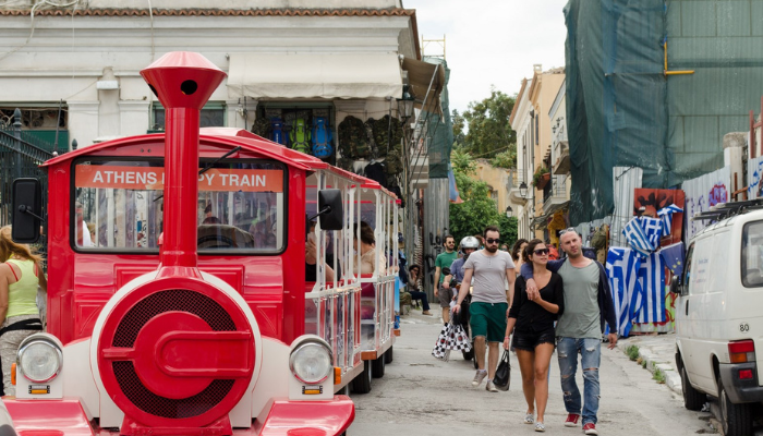 Red Athens sightseeing train driving through Plaka district with tourists walking nearby.