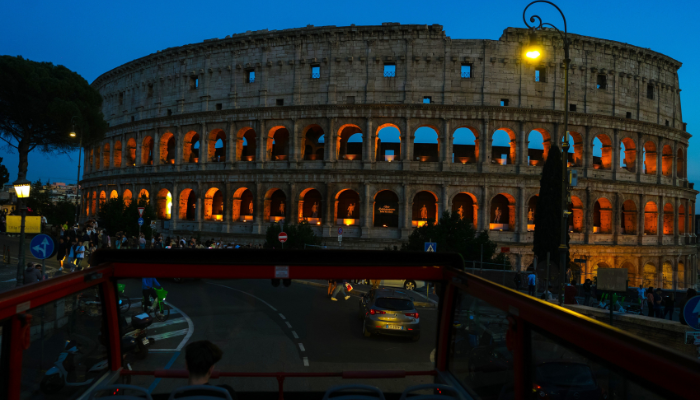 The Colosseum illuminated at night with glowing arches, viewed from an open-top sightseeing bus in Rome.