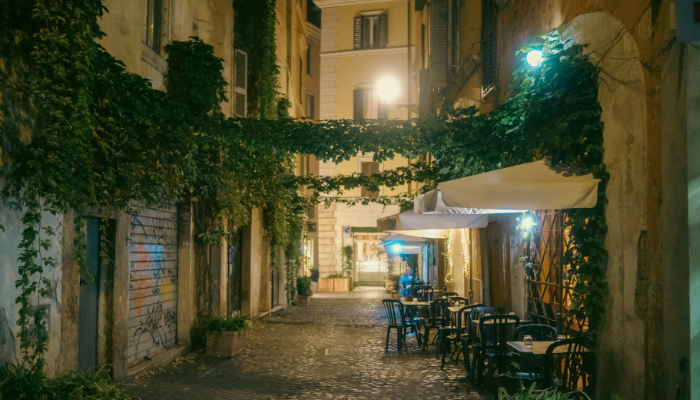 Quiet cobblestone street in Trastevere, Rome at night with ivy-covered walls and outdoor café tables.