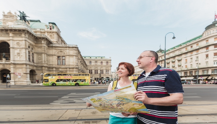 Tourists standing near the Vienna State Opera with a city map while a sightseeing bus passes behind them.