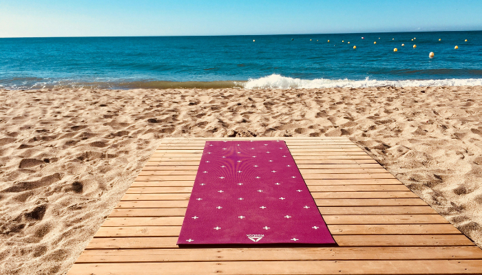 A yoga mat lies on a sunny sandy beach along Spain’s Costa del Sol coastline with clear Mediterranean water.