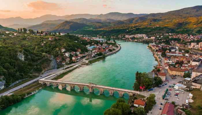 River flowing through Bosnia town with arched stone bridge, green water, and hills in the background.
