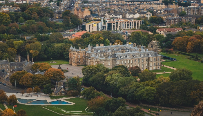 Aerial view of Holyrood Palace surrounded by green parkland and Edinburgh cityscape.