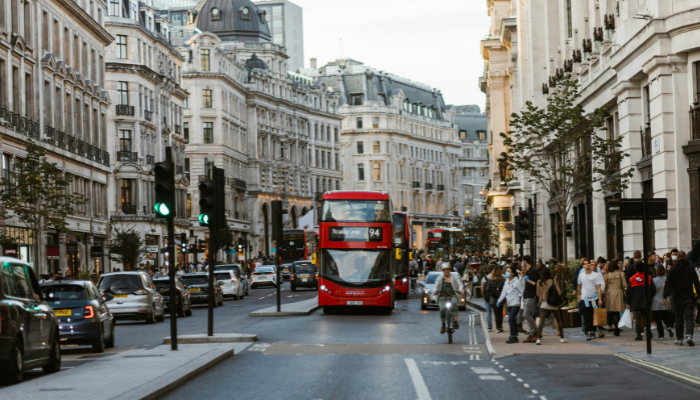 Red double-decker bus driving through a busy London street surrounded by historic buildings and pedestrians.