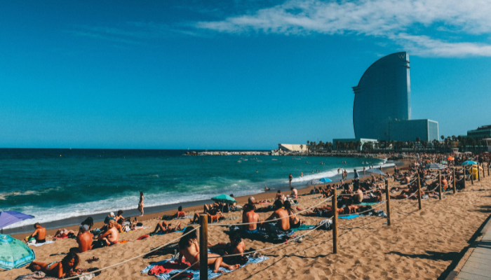 People relaxing on the sandy shores of Barceloneta Beach with the W Barcelona Hotel visible along the Mediterranean coastline.