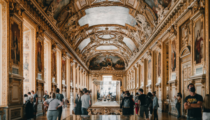 Visitors exploring the grand gallery inside the Louvre Museum, surrounded by ornate ceilings, classical paintings and historic architecture.